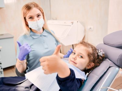 child in a dentist chair giving a thumbs up and happy, the dentist is behind her giving a thumbs up also.
