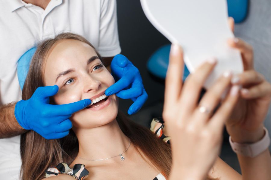 woman in a dentist chair, she is looking at her teeth in a mirror, the dentist is behind her pointing at her teeth