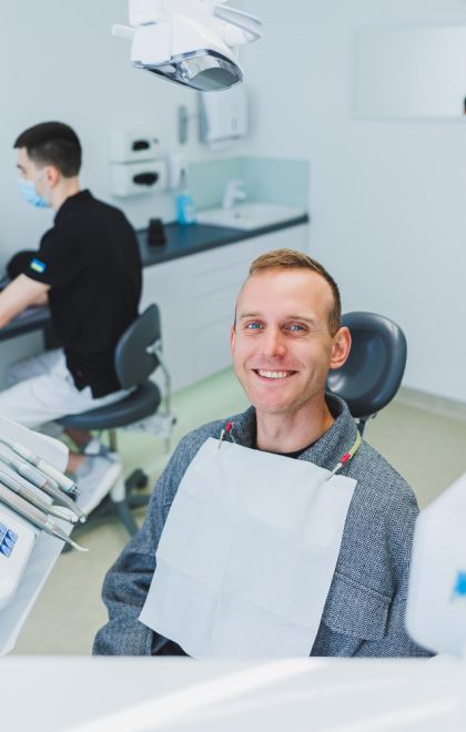 A young man is sitting in a dental chair and a dentist is working at a computer desk